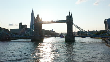 Low-angle-view-of-Tower-bridge-and-Shard-skyscraper-against-sun.-Low-flight-above-River-Thames-water-surface.-London,-UK