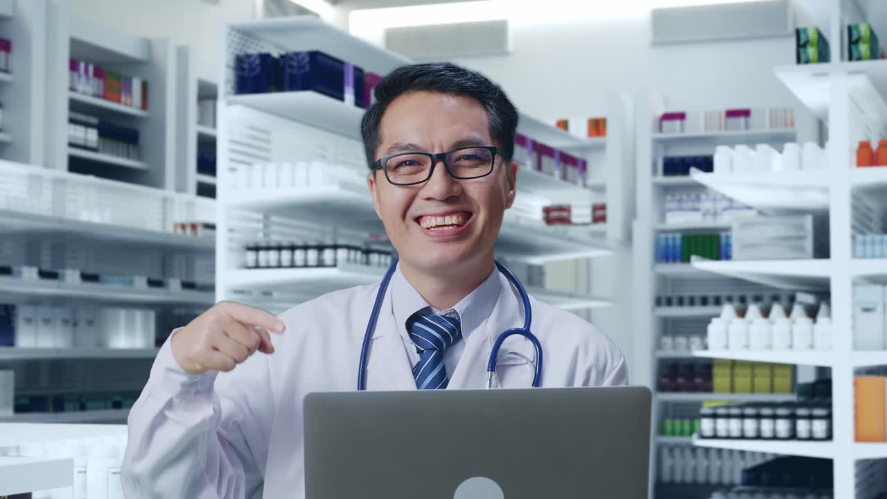 스테토스코프를 들고 있는 아시아 남성 의사가 약국에 서서 손에 노트북을 가리키며 웃고 있다.