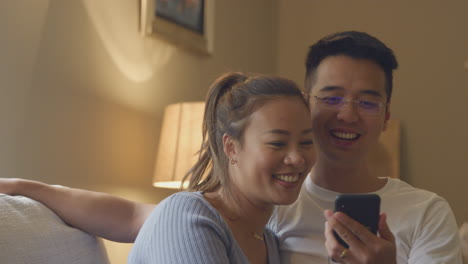Lächelndes-Asiatisches-Paar-Entspannt-Sich-Zu-Hause-Auf-Dem-Sofa-Und-Schaut-Abends-Gemeinsam-Auf-Das-Mobiltelefon