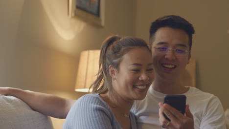 Una-Pareja-Asiática-Sonriente-Relajándose-En-El-Sofá-De-Casa-Mirando-El-Teléfono-Móvil-Juntos-Por-La-Noche