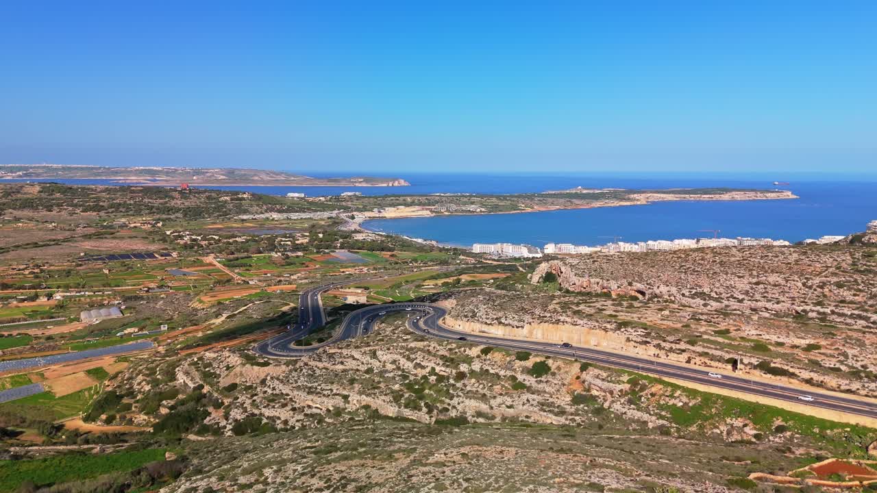Vista aerea con drone della baia di Għadira a Mellieħa, Malta, che evidenzia la costa mediterranea, strade tortuose, campi agricoli e il vibrante contrasto tra terra e mare sotto un cielo azzurro e limpido.