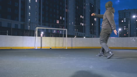 el jugador golpea la pelota de fútbol con potencia durante la sesión de entrenamiento nocturna en el campo al aire libre, la pelota rebota en el poste de la portería bajo las luces brillantes del estadio, rodeada por el paisaje nocturno urbano