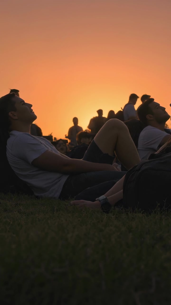 Video verticale: Uomo sdraiato sull'erba al tramonto, il sole che cala mentre un escursionista con zaino passa.