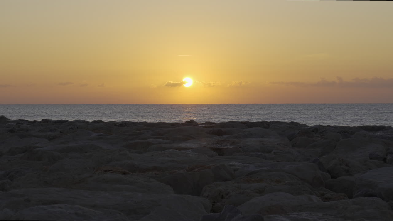 저속 촬영은 바다 위의 일출을 포착하여 물에 반사되고 바위가 많은 전경을 비춥니다.
