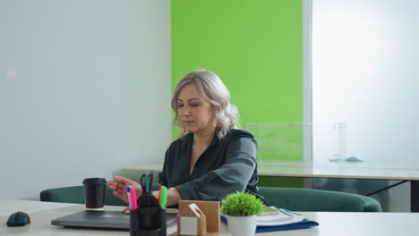 realtor in dark blouse adjusts smartphone and documents on desk, preparing workspace before opening laptop to begin work in bright modern office with vibrant green wall and minimal decor
