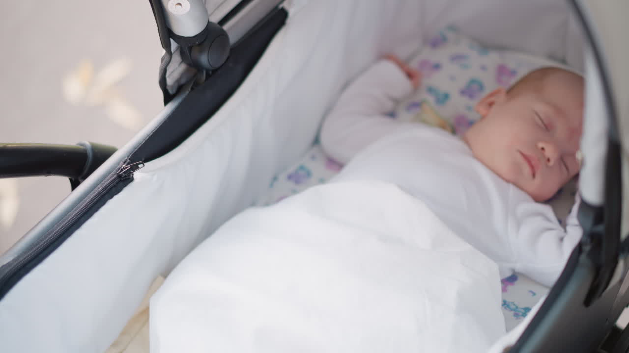 Neonato che dorme nella navicella del passeggino, primo piano intimo di un neonato avvolto in una coperta bianca, una mano delicata sistema la coperta, luce soffusa e atmosfera accogliente, stile di vita materno tranquillo che suggerisce la genitorialità.
