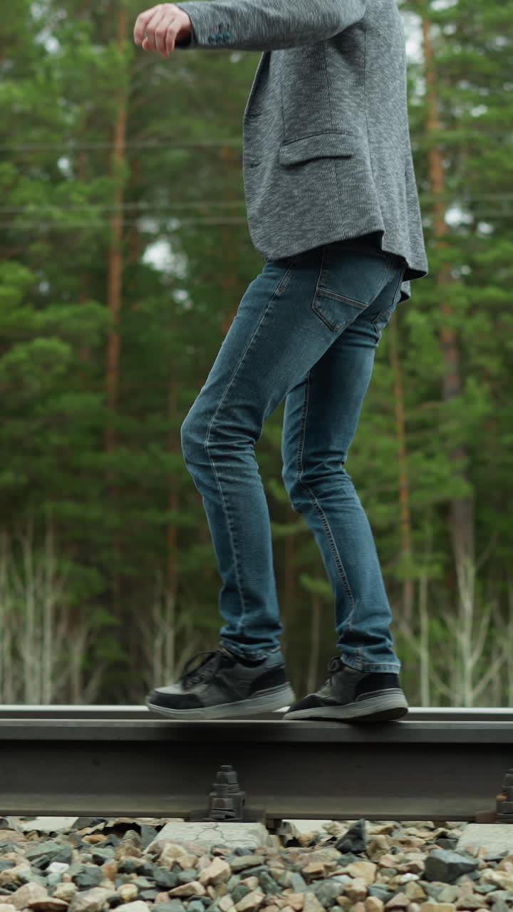 회색 슈트 재과 파란 청바지를 입은 남자가 철도 트랙을 따라 조심스럽게 고, 균형을 맞추고, 배경에는 울창한 초록색 나무가 있습니다.