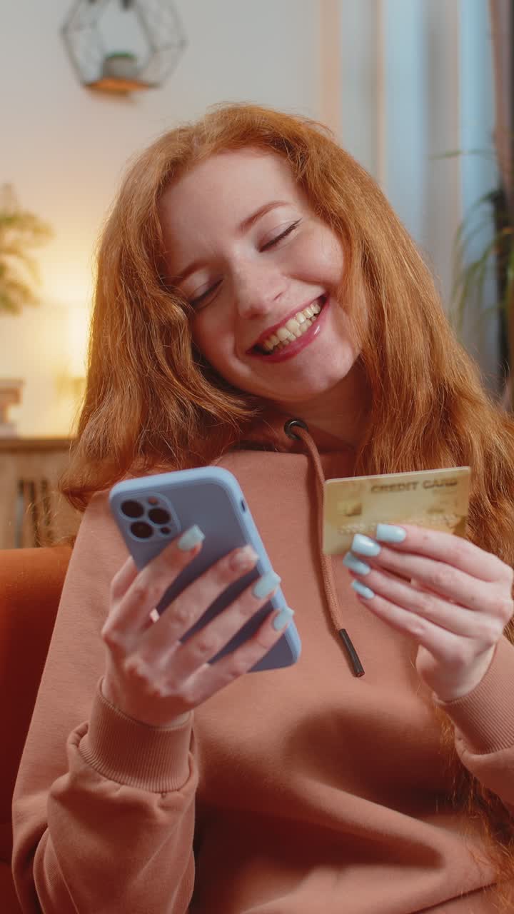 joven feliz usando tarjeta de crédito y teléfono inteligente mientras hace compras en línea sentada en el sofá