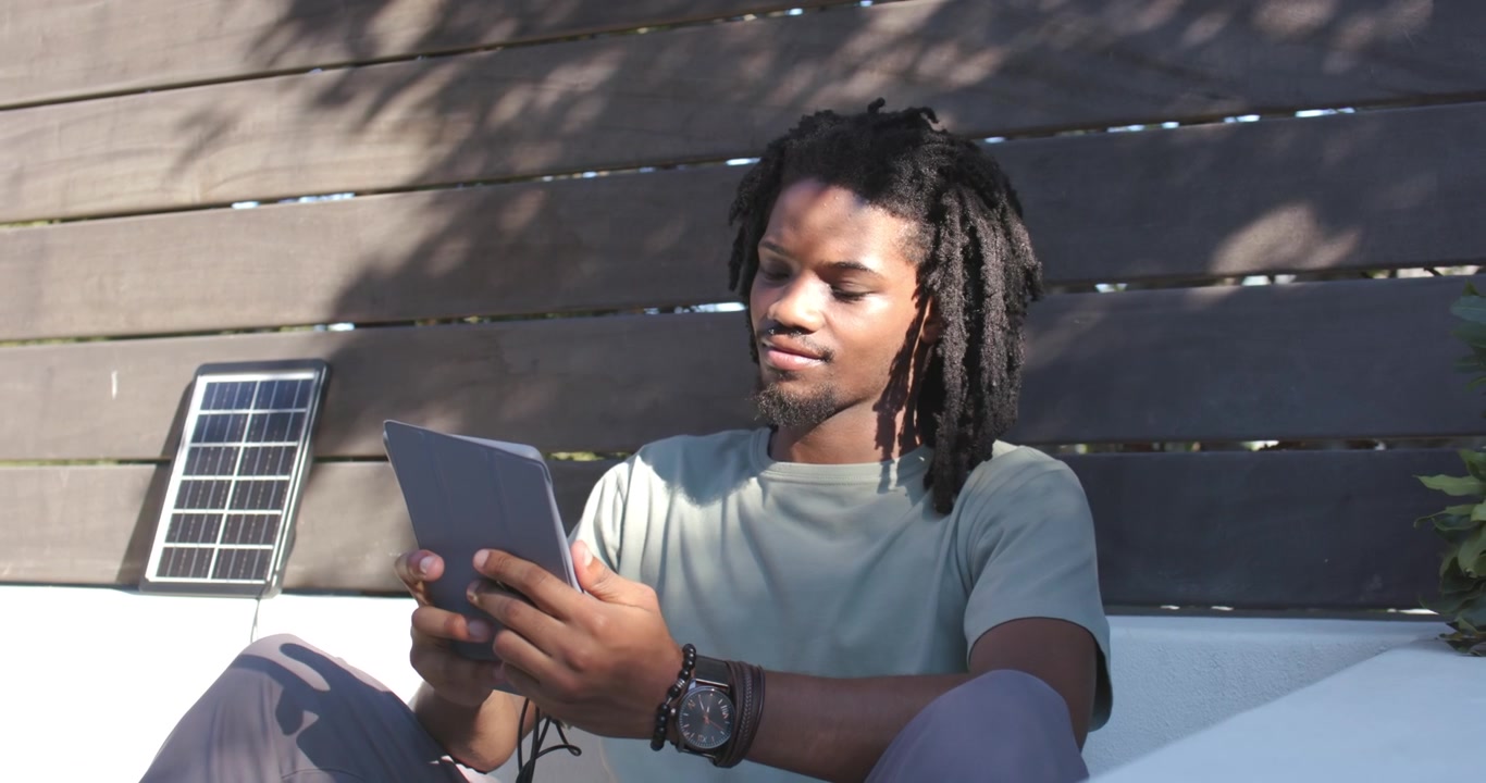 African American man sitting on sofa holding tablet tilting to reduce glare, solar panel left