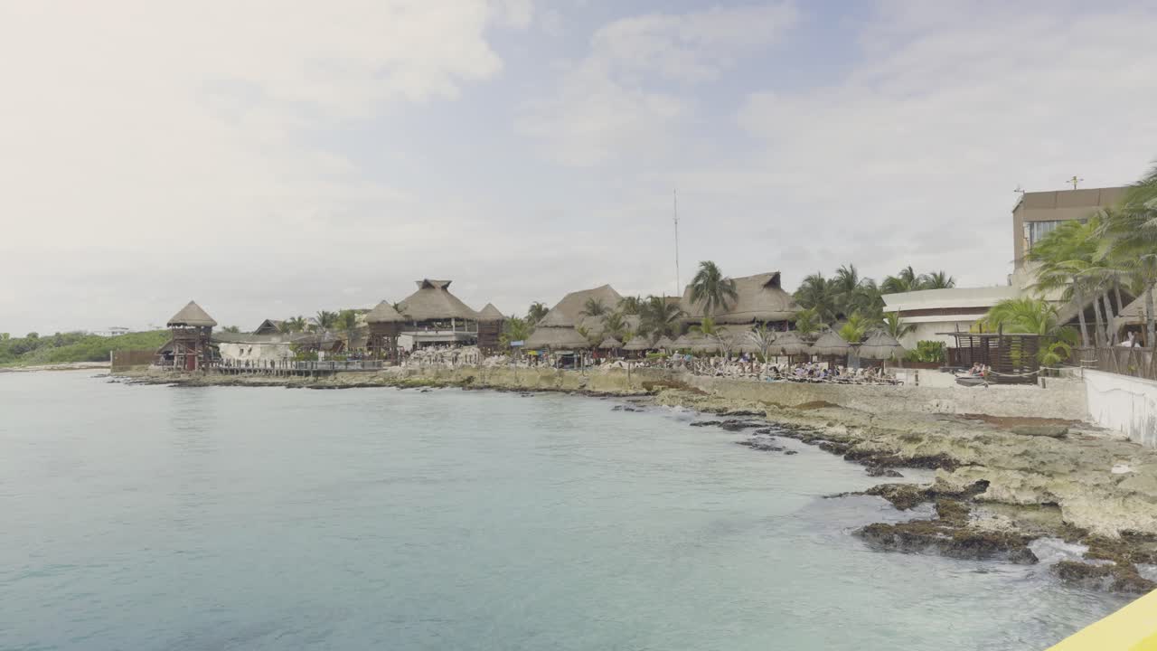 View from the Costa Maya cruise ship dock, with gentle waves crashing on rocks, people walking toward the port center, and an overcast blue sky above. A serene, vibrant coastal scene.