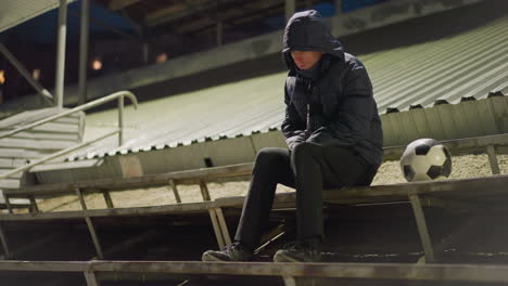 a man wearing jacket sits alone in a dimly lit stadium, with his head bowed down and hands clasped between his legs, quietly contemplating, with a soccer ball placed beside him