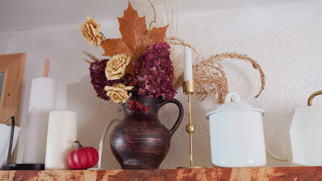 Neatly arranged flower vessel on rustic wooden shelf with dried floral bouquet, white ceramic pots, golden candle stand, paper towel roll, and miniature red pumpkin in cozy well-lit kitchen