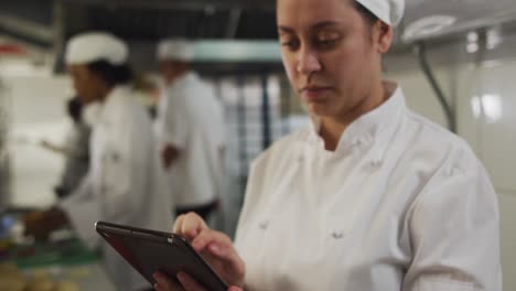 chef caucásica usando una tableta en la cocina de un restaurante con otros chefs en el fondo