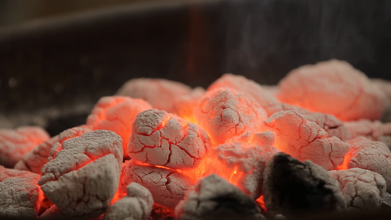 Braci ardenti di carbone in un barbecue, primo piano di carboni incandescenti coperti di cenere bianca, con leggero fumo che sale.
