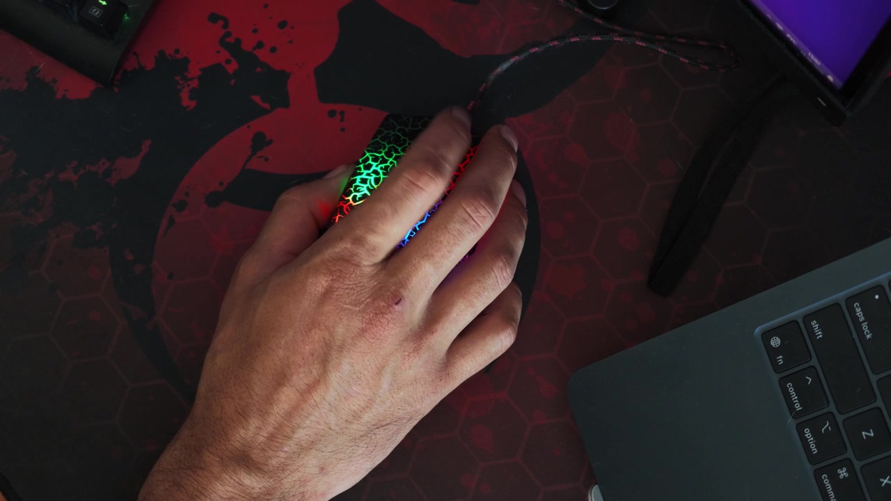 Una mano afferra un mouse da gaming illuminato a colori arcobaleno su un tappetino esagonale, con illuminazione RGB che cambia colore mentre la mano effettua leggere regolazioni, e una tastiera di laptop è parzialmente visibile nelle vicinanze.