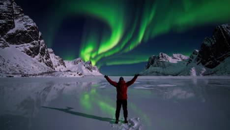 person under the vibrant northern lights over snowy mountains and a frozen lake
