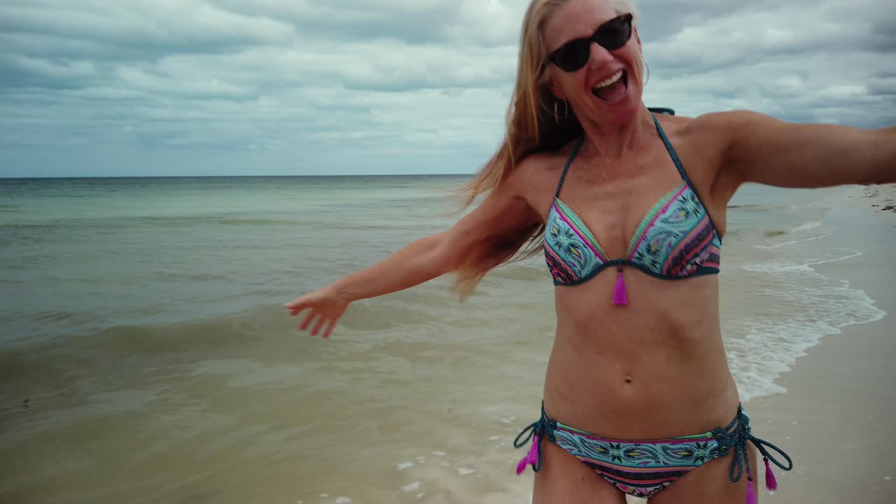 Ripresa da dietro di una giocosa donna bionda matura in occhiali da sole e bikini che corre lungo una spiaggia e guarda indietro verso la telecamera con il braccio teso verso qualcuno.