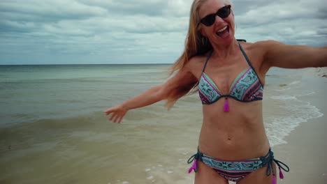 Rear-shot-of-playful-blonde-mature-woman-in-sunglasses-and-bikini-running-along-a-beach-and-looking-back-at-the-camera-with-arm-back-reaching-out-to-someone