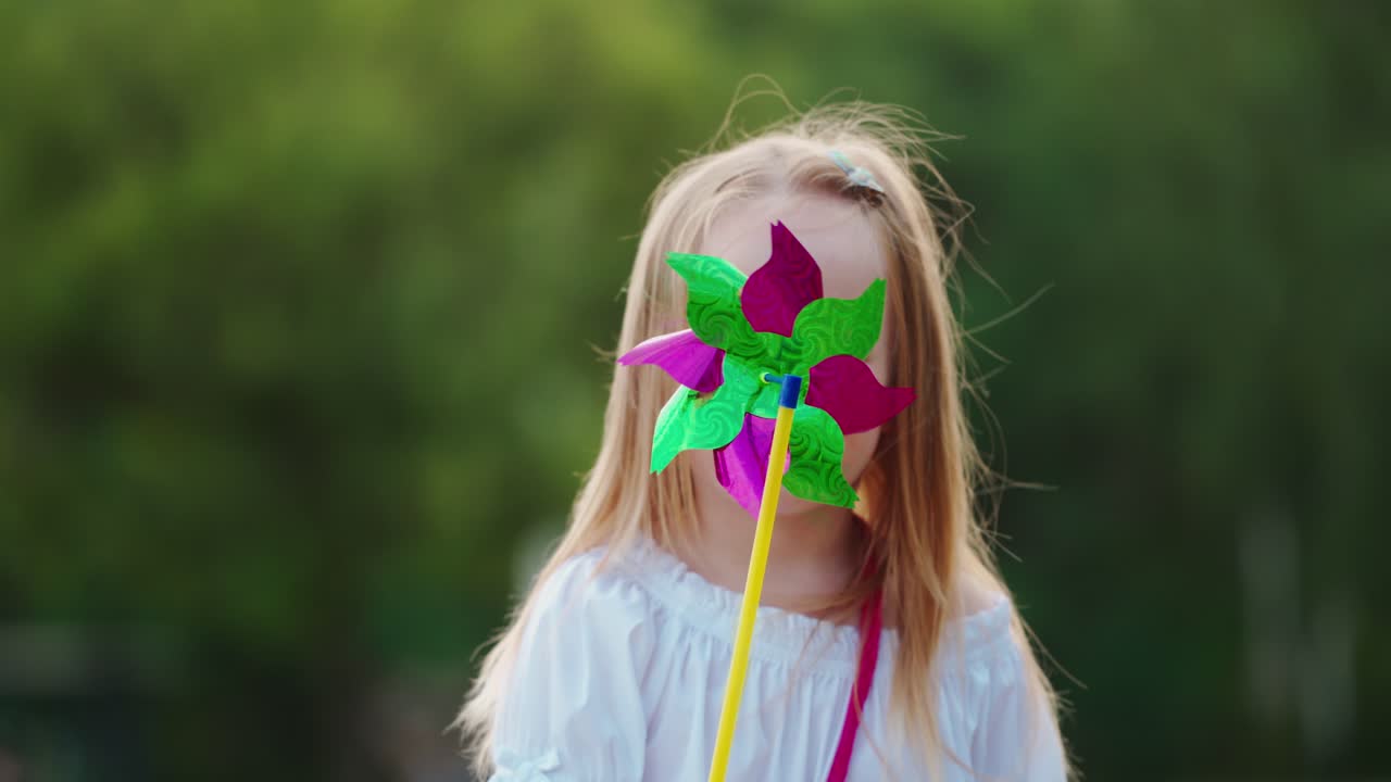 Portrait of a little girl with a pinwheel. Positive child hiding her face by a toy windmill and smiling at camera outdoors.