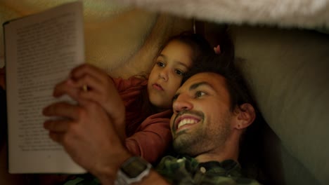 a little brunette girl in a pink dress lies in bed with her father who tells her stories, they read a book together and look at pictures with a flashlight on near a gray sofa at home