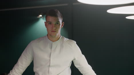 young man in white shirt with serious facial expression stands in dimly lit background. focused gaze and intense mood create a dramatic atmosphere, illuminated by overhead lighting in dark setting