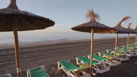 panorámica lenta a la izquierda en la playa de marbella al amanecer con tumbonas y sombrillas de paja, pequeñas olas tranquilas en una playa vacía, inspiración de viajes de lujo de ensueño