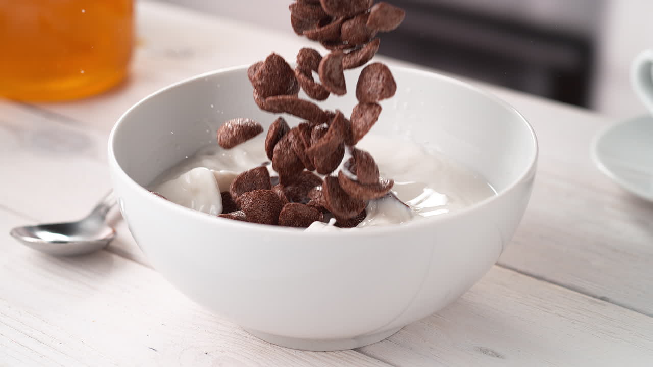 カカオの状の穀物がキッチンテーブルの上にある白いミルクの鉢にスローモーションで落ちている