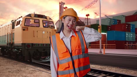 asian female engineer with safety helmet smiling to camera and making call me gesture with freight cargo train at port