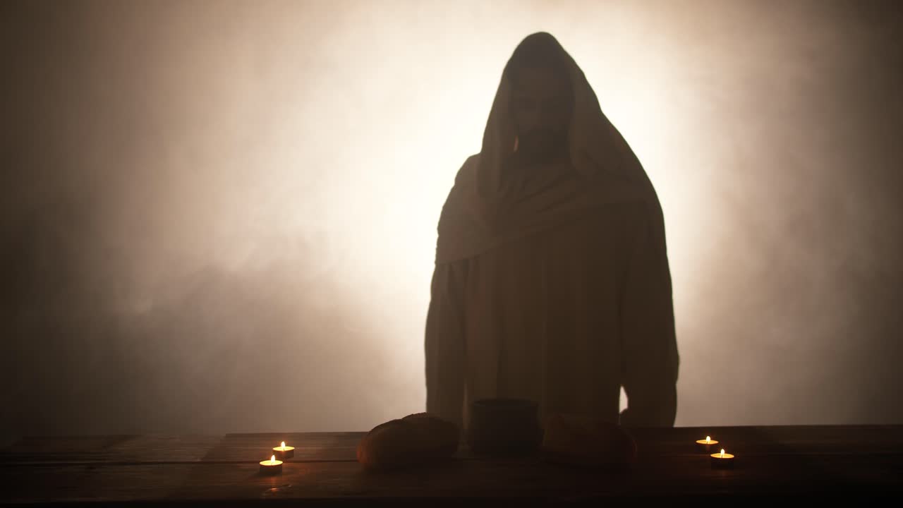 The silhouette of Jesus Christ standing near a table with Christian sacraments of bread and wine for last supper, communion, Easter symbolism. In cinematic slow motion with ambient light.