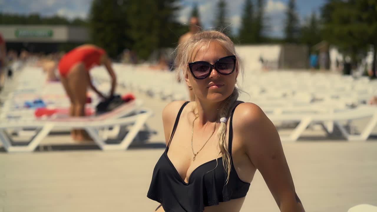 Primo piano: Ragazza bionda seduta su una sdraio in spiaggia con occhiali da sole, ha i capelli intrecciati in una treccia e si guarda intorno.