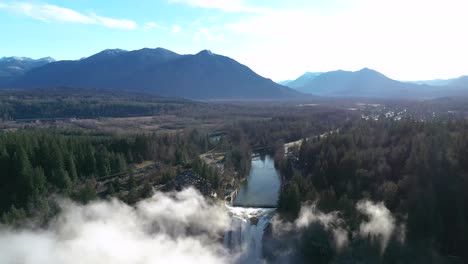 snoqualmie falls behind clouds surrounded with trees in park and mountain in background in washington, usa