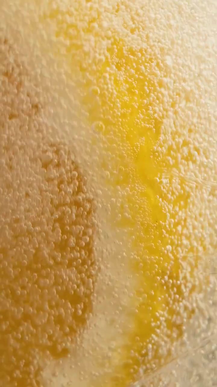 Primo piano macro cinematografico verticale in super slow-motion di una fetta d'arancia all'interno di un bicchiere da cocktail. Viene versata una bevanda gassata, facendo fluttuare dinamicamente bollicine scintillanti sul frutto agrumato