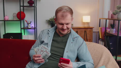 Smiling-happy-young-man-counting-money-cash-and-use-mobile-phone-calculate-domestic-bills-at-home