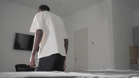 young man moving swiftly through modern bedroom, walking toward television, black shorts, white t-shirt, minimalistic interior, modern design, energetic morning movement, start of day, active