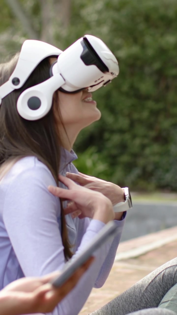 Video verticale: Donna si sistema il visore VR, in top, guarda in alto sorridendo su una panchina del parco mentre una mano tocca il cellulare.