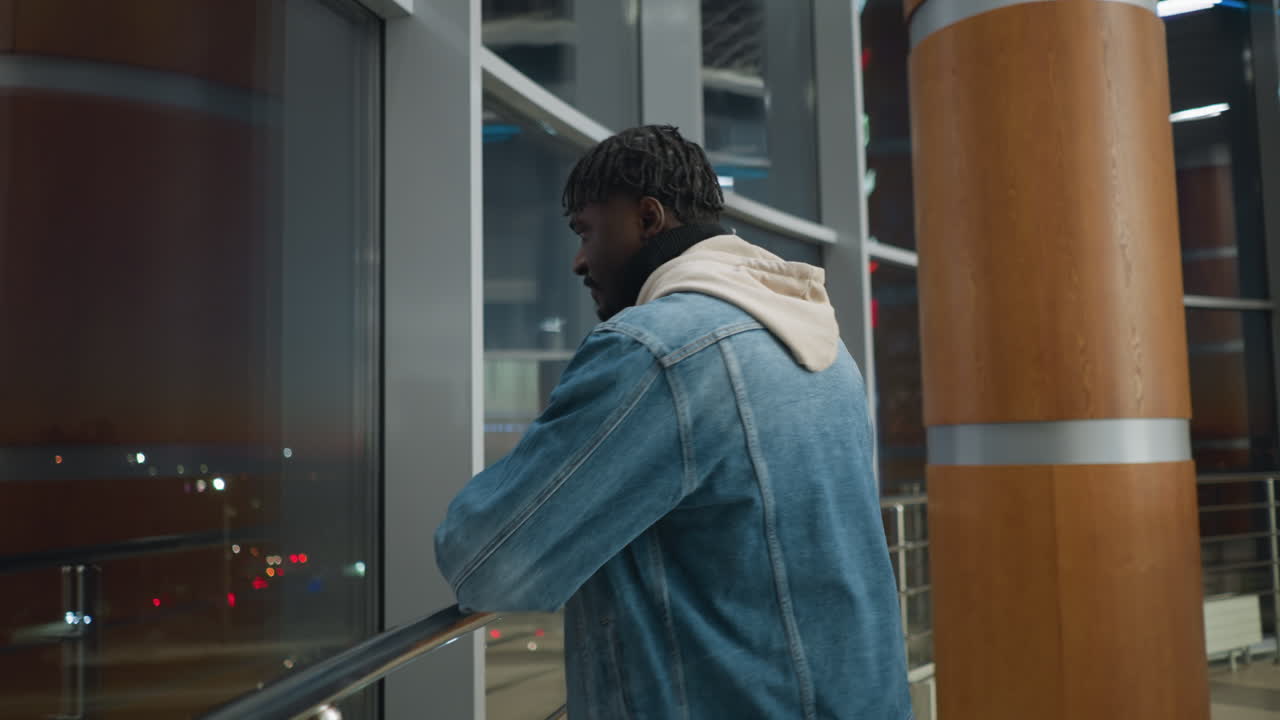 Young man in denim jacket and hoodie walking alone in modern building near large windows at night, viewed from behind, reflecting calm atmosphere with city lights visible through glass panels