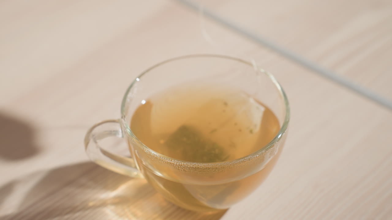 Close-up of tea bag floating inside a clear cup with freshly brewed tea. Tea is steeping, with steam rising from the cup, captured in a calm kitchen setting, preparing hot beverage