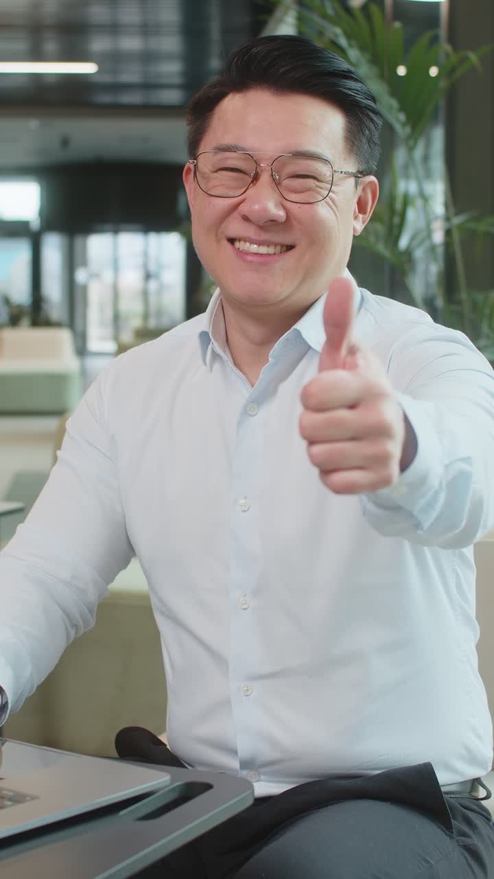 Happy young businessman working on laptop looking at camera showing thumbs up in modern office lobby