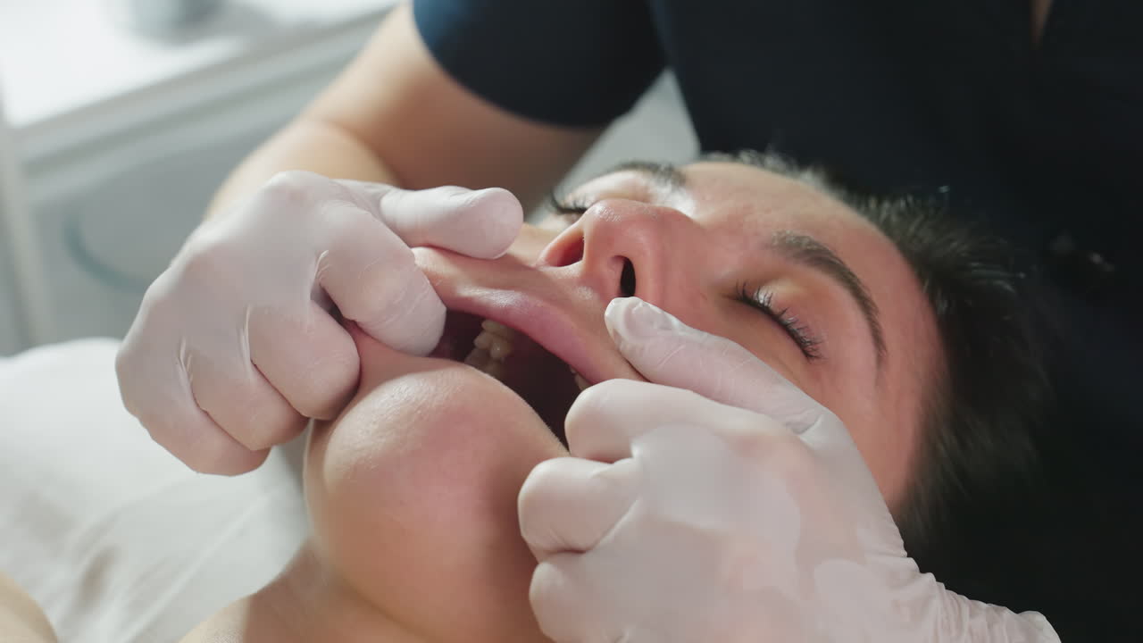Close up of recipient lying on spa bed eyes closed lips gently parted as beauty expert in gloves dips fingertips into mouth massaging lips and jawline for deep facial relaxation and care