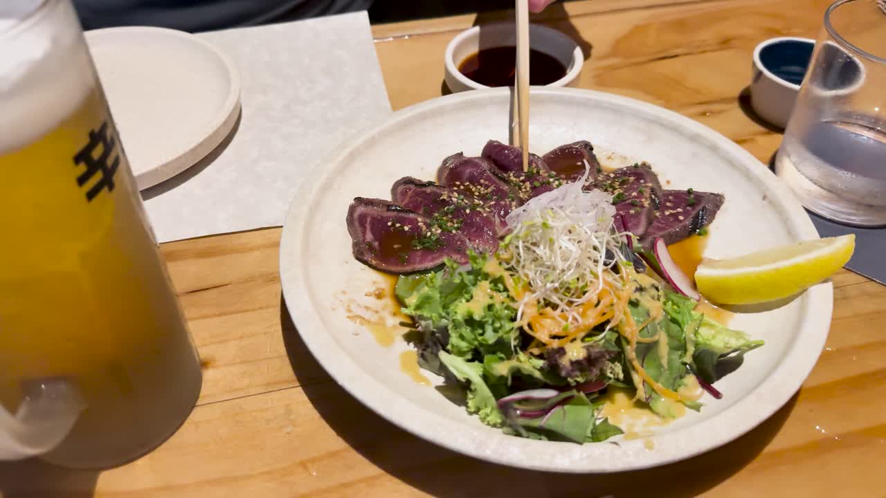 Wagyu beef tataki served with salad and lemon slice on a wooden table, accompanied by drinks in a Japanese restaurant setting