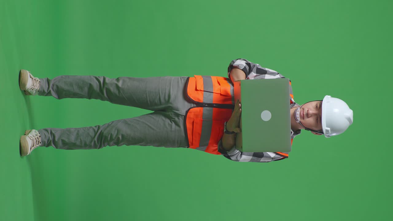 Full Body Of Asian Male Engineer With Safety Helmet Working On A Laptop And Looking Around While Standing In The Green Screen Background Studio