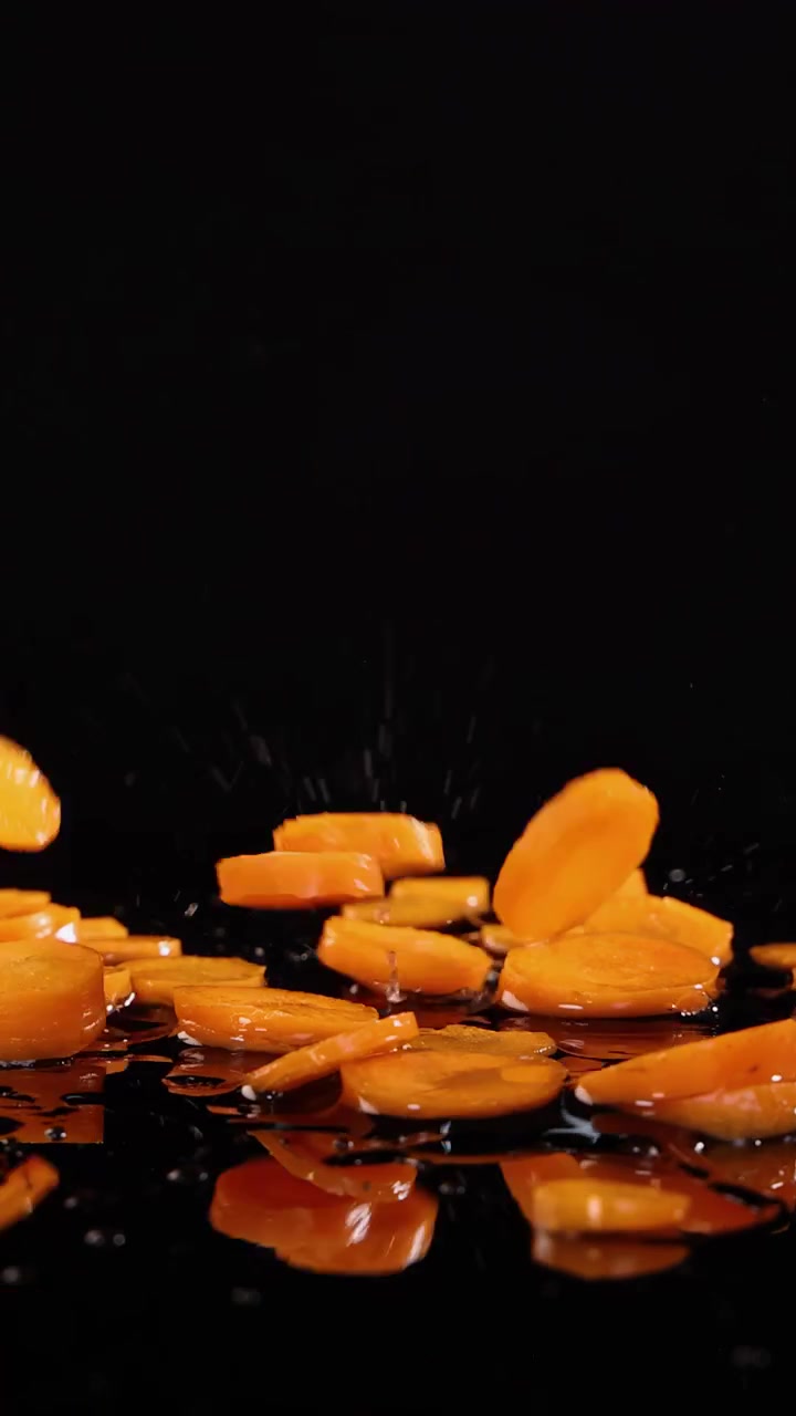Carote baby affettate cadono e schizzano nell'acqua su uno sfondo nero, riprese in slow motion ad alta velocità con illuminazione drammatica e riflessi nitidi.