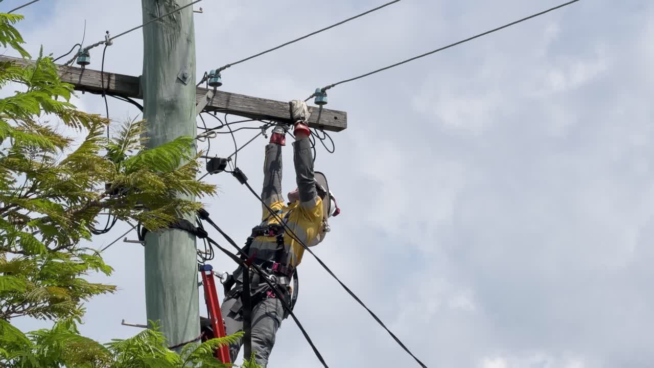 Utility worker repairs overhead power cables on pole, using safety gear, in daylight, wide shot