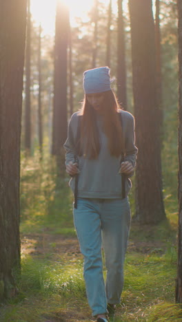 mujer joven caminando a través de un bosque sereno mirando a su alrededor, sosteniendo correas de mochila como la suave luz del sol de la tarde crea un brillo suave a su redor, árboles altos y vegetación exuberante enmarcan su camino
