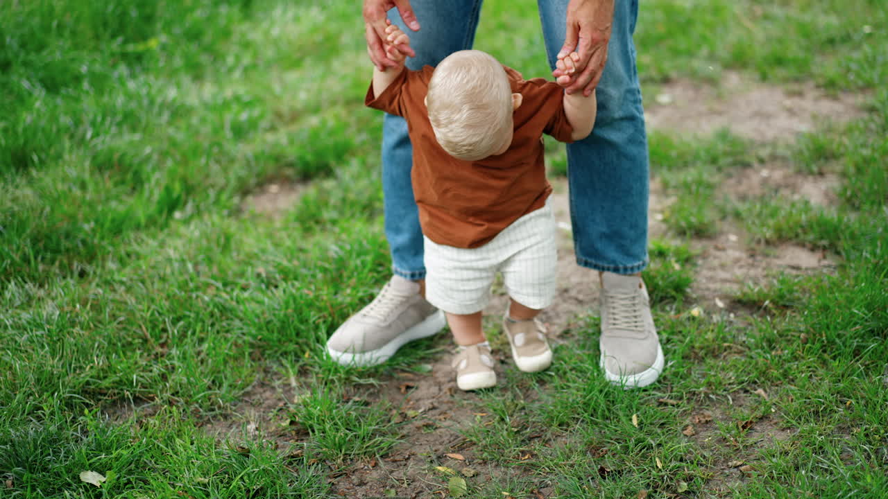 Unrecognized father wearing jeans leads his little son. Lovely blond baby is learning to walk in the nature.