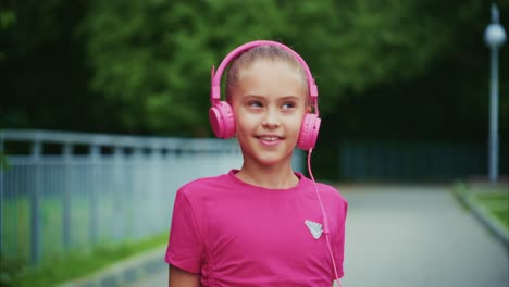a joyful girl enjoys her music while walking on a pathway, showcasing her pink headphones and bright attire in a lush green environment