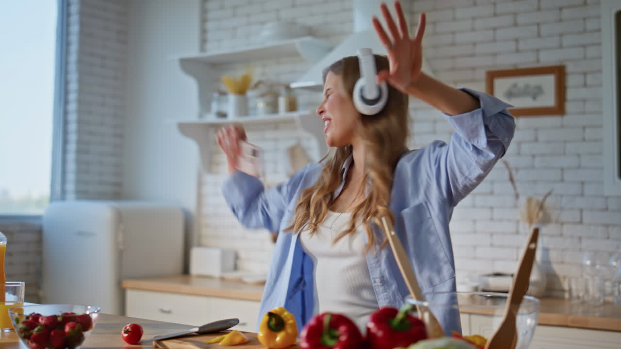 Joyful woman using mobile phone in kitchen while cooking. Lady listening music