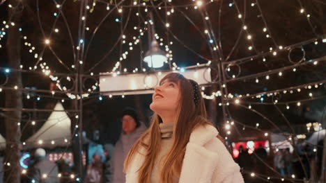 Hermosa-Joven-Con-Cabello-Oscuro-Y-Bata-Blanca-Sonriendo-Directamente-A-La-Cámara-Y-Mirando-La-Decoración-De-Luces-Navideñas-En-Un-Mercado-Navideño-Durante-Una-Fría-Tarde-De-Invierno-En-Cámara-Lenta