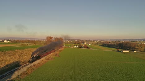 Luftaufnahme-Einer-Antiken-Dampflokomotive,-Die-Sich-Während-Der-Goldenen-Stunde-Am-Späten-Nachmittag-Mit-Personenwagen-Nähert-Und-Rauch-Und-Dampf-Bläst