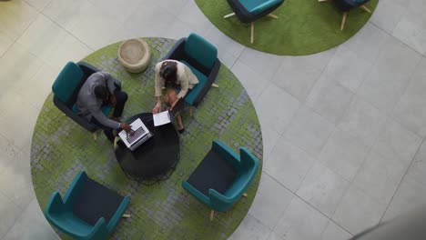 Diverse-businessman-and-businesswoman-using-laptop-in-meeting-in-lobby-of-modern-office